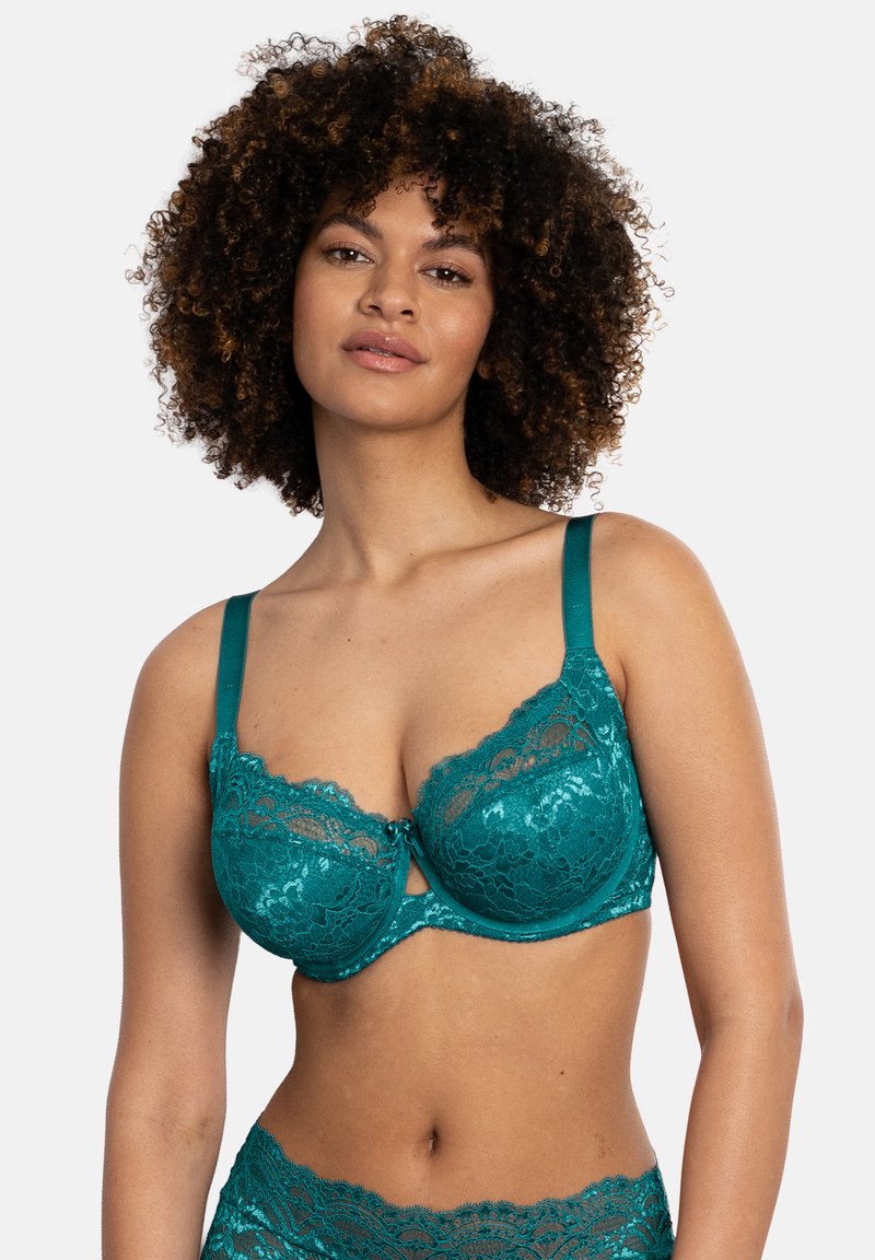 Frau mit lockigem Haar, die einen türkisen Spitzen-BH und passende Spitzen-Unterwäsche mit hoher Taille trägt, steht vor einem einfarbigen hellen Hintergrund.