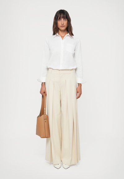 Chemise blanche à boutons avec une poche poitrine, associée à un pantalon beige plissé taille haute et des chaussures crèmes à bouts pointus. Tient un sac à épaule beige.
