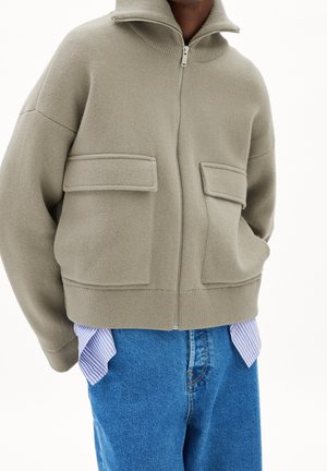 Person trägt eine übergroße beige Zip-up-Jacke mit großen Fronttaschen, blaue Jeans und ein gestreiftes Shirt, das am Saum hervorlugt.