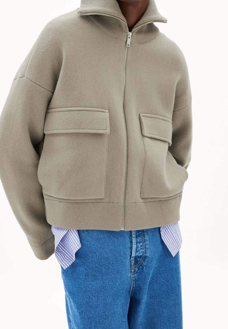 Personne portant une veste zippée beige oversize avec de grandes poches avant, un jean bleu et une chemise rayée qui dépasse de l’ourlet.