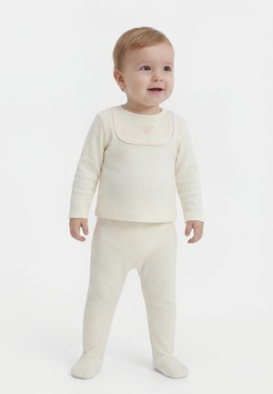 Tipegő áll, krémszínű bordás hosszú ujjú felsőt és hozzáillő lábfejes nadrágot visel, mosolyog, és kissé jobbra néz.