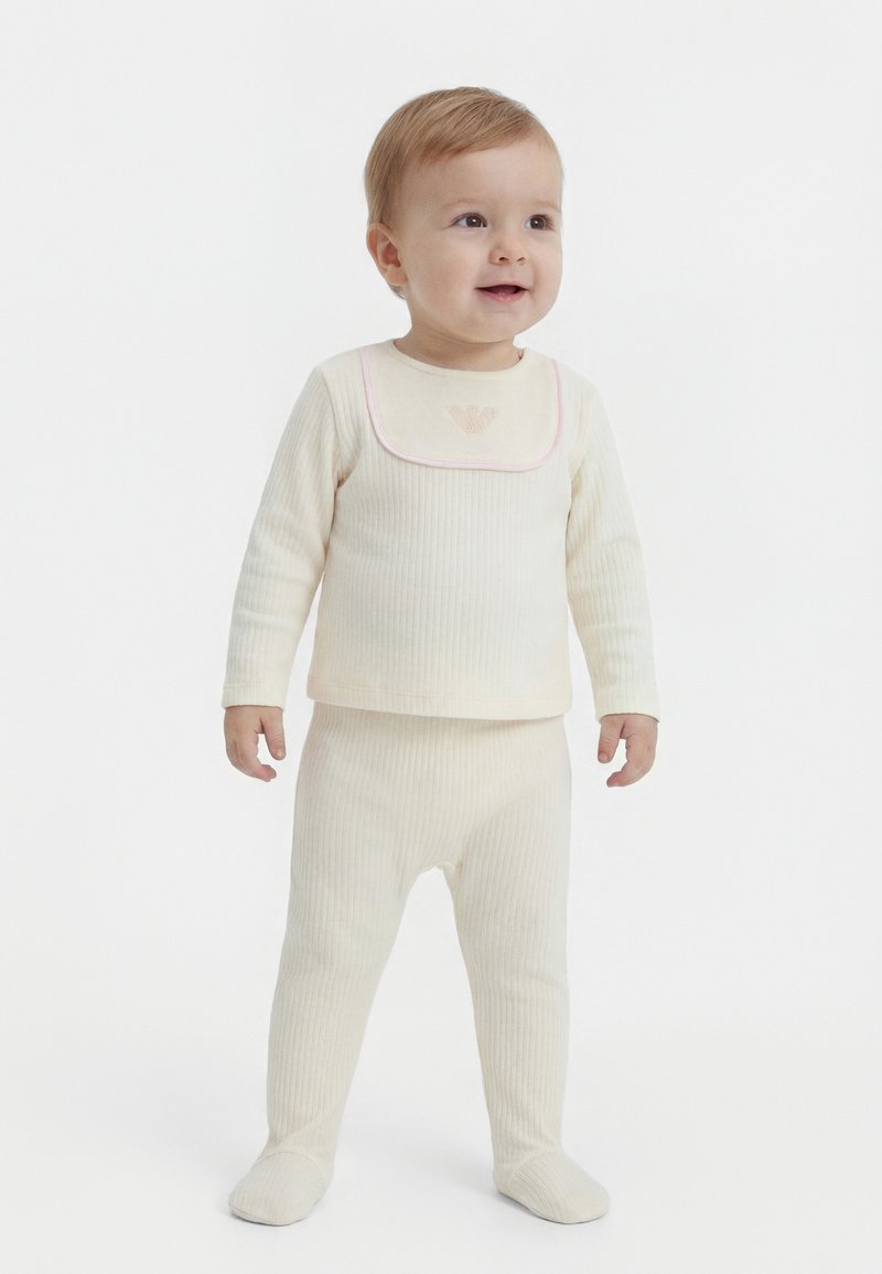 Väikelaps seisab, seljas helekreemjas soonikuga pikad varrukad ja sobivad jalaosaga püksid, naeratab ja vaatab kergelt paremale.