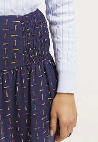 Marine rok met een geplooide taille, voorzien van een print van kleurrijke lippenstiften. De stof is lichtgewicht met een zachte textuur. Lichte blauwe kabelgebreide trui mouwen zichtbaar.