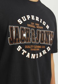 Sort bomulds t-shirt med et stort grafisk print i hvid og orange: "JACK & JONES," med tekstelementer og etableringsdato.