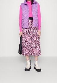 Doudoune violette matelassée, haut à manches longues rose, jupe midi fleurie en rose et noir, bottines blanches avec accents noirs, portant un sac noir.
