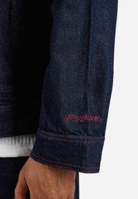 Donkerblauwe spijkerjack met een mouw die is voorzien van een rood geborduurd "PEGADOR" logo en contrasterende stiksels. De textuur lijkt soepel met een gestructureerde pasvorm.