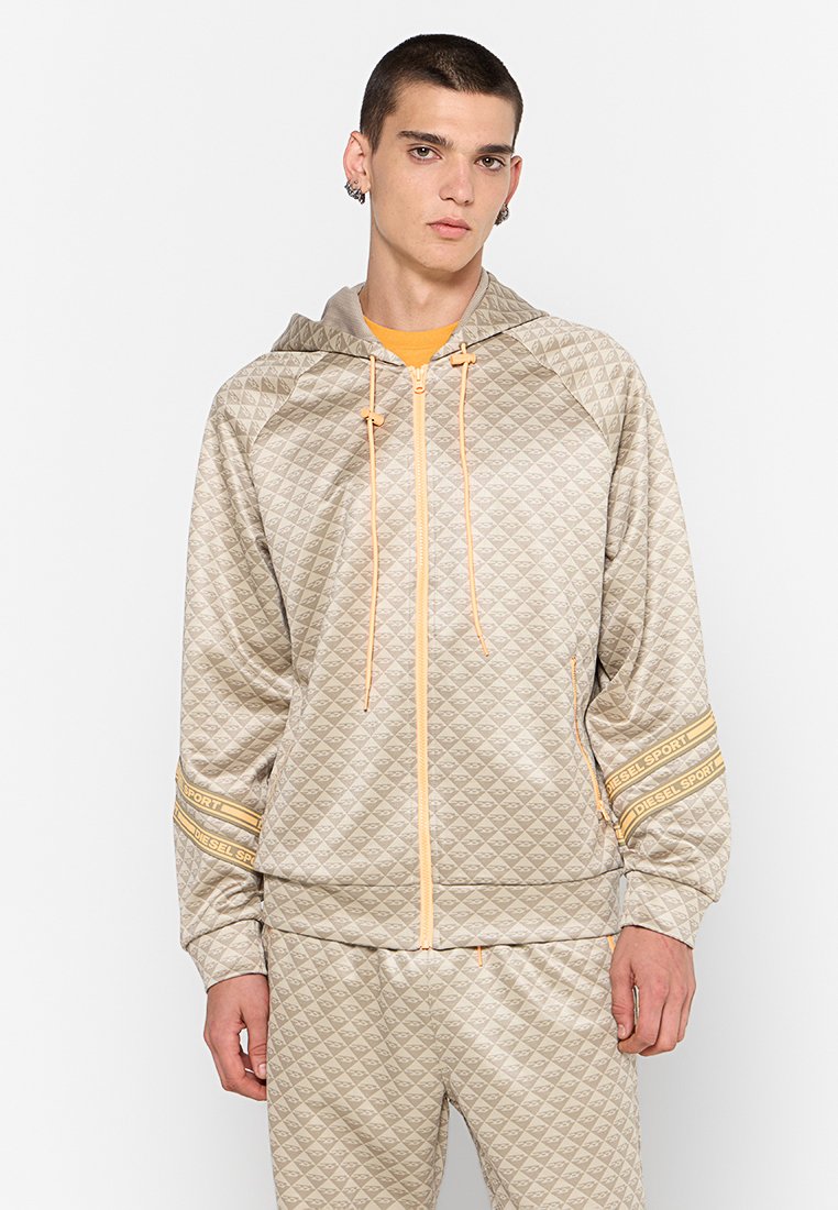 Giacca beige imbottita con chiusura a zip e cappuccio, con dettagli arancioni e strisce brandizzate, abbinata a pantaloni coordinati, realizzati in tessuto liscio.