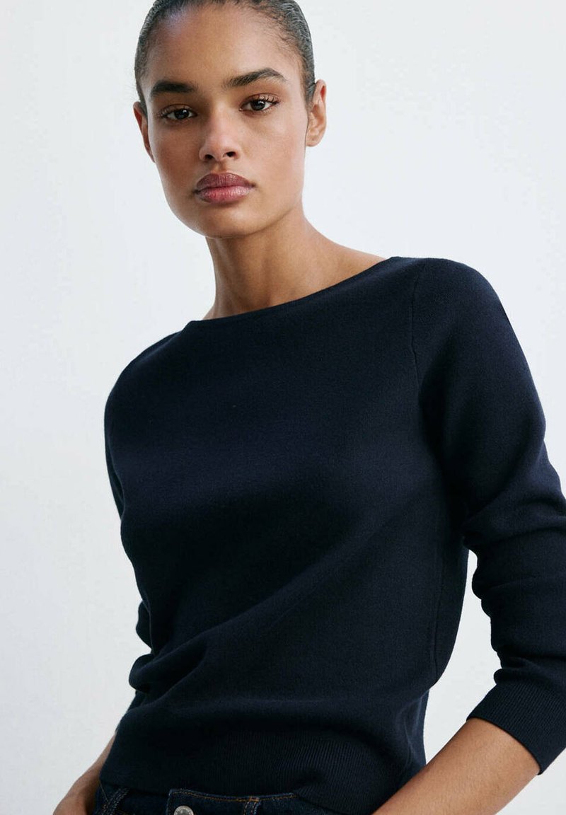 Femme aux cheveux coiffés en arrière portant un pull noir ajusté à manches longues sur un fond clair uni.