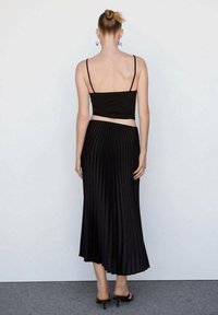 Top crop nero abbinato a una gonna maxi nera plissettata. La gonna presenta un design fluido con pieghe verticali. Sandali neri con tacco.