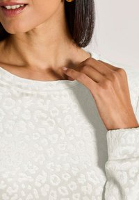 Frau mit Goldohrringe in Form von Ringen, die einen strukturierten weißen Leopardenmuster-Oberteil trägt und sanft mit der rechten Hand das Schlüsselbein berührt.