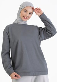 Sudadera gris con cuello redondo y mangas largas, hecha de material suave. El modelo lleva un pañuelo de cabeza de color gris claro. Fondo limpio.