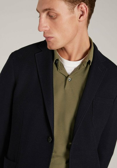 Blazer blu navale con texture e un bottone, abbinato a una polo verde oliva con colletto e tre bottoni.