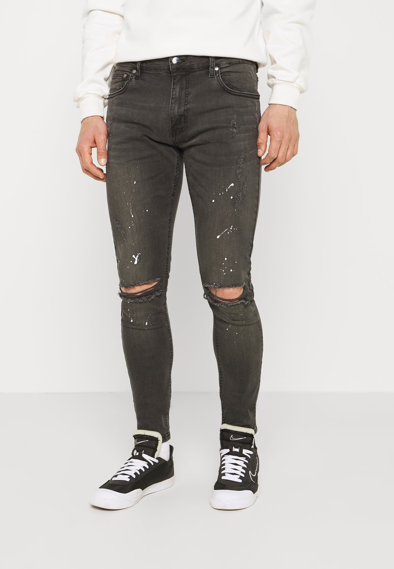 Pantalones vaqueros ajustados negros con roturas desgastadas en las rodillas, detalles de salpicaduras de pintura blanca y un diseño tradicional de cinco bolsillos, combinados con zapatillas negras.