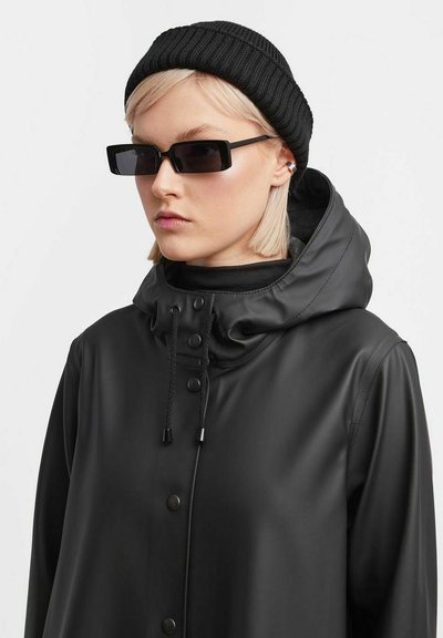 Zwarte waterdichte hooded jas met knopen aan de voorkant, trekkoord bij de hals, textuurmateriaal, gedragen met een zwarte gebreide beanie en hoekige zonnebril.