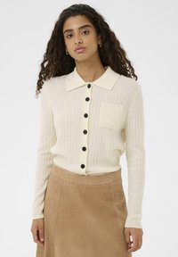 Krēmkrāsas ribota adīta kardigāna ar asas formas apkakli, melni pogas priekšpusē un kreisajā krūtīs kabatu. Gluda tekstūra.