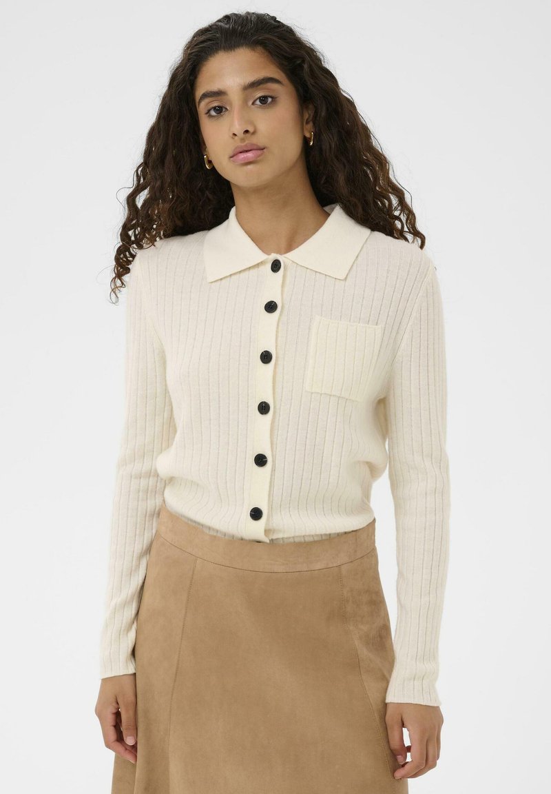 Krēmkrāsas ribota adīta kardigāna ar asas formas apkakli, melni pogas priekšpusē un kreisajā krūtīs kabatu. Gluda tekstūra.