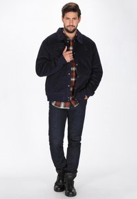 Marine blauwe corduroy jas met een gewatteerde kraag, knoopsluiting en voorzakken, gelaagd over een geruit overhemd en donkere jeans, gecombineerd met zwarte laarzen.