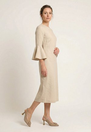 Frau trägt ein beiges Midikleid mit ausgestellten Ärmeln und passenden beigen Slingback-Pumps mit Absatz, steht vor einem schlichten hellen Hintergrund.