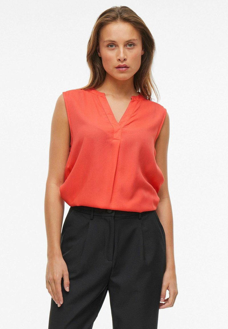 Blusa rossa senza maniche con scollatura a V e una leggera piega sul davanti, abbinata a pantaloni neri su misura. Tessuto liscio e vestibilità rilassata.