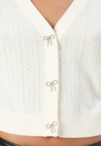 Gilet en tricot blanc avec un col en V, présentant un motif en diamant, surmonté de trois accents en forme de nœuds ornés de bijoux le long de la ligne de boutons.