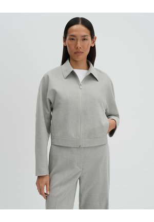 Frau mit glattem dunklem Haar, die eine hellgraue Jacke mit Reißverschluss und passende Hose trägt, steht vor einem schlichten hellen Hintergrund.