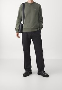 Felpa verde oliva con collo rotondo e piccolo logo, abbinata a pantaloni neri larghi e scarpe nere, mentre tiene una borsa a spalla nera.