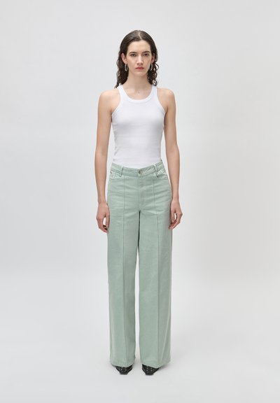 Γυναίκα με μακριά σκούρα μαλλιά φοράει λευκό αμάνικο τοπ, ανοιχτό πράσινο παντελόνι με ψηλή μέση και φαρδιά μπατζάκια, και σκουλαρίκια κρίκους, στέκεται μπροστά σε απλό φόντο.