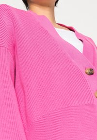 Cardigan rosa a V con texture a coste, dotato di bottoni in legno. Il design presenta maniche ampie e una vestibilità rilassata.