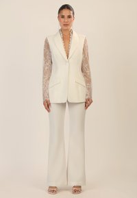 White tailored vest with lace long sleeves, deep V-neck, and scalloped collar; paired with white flared trousers and strappy sandals.