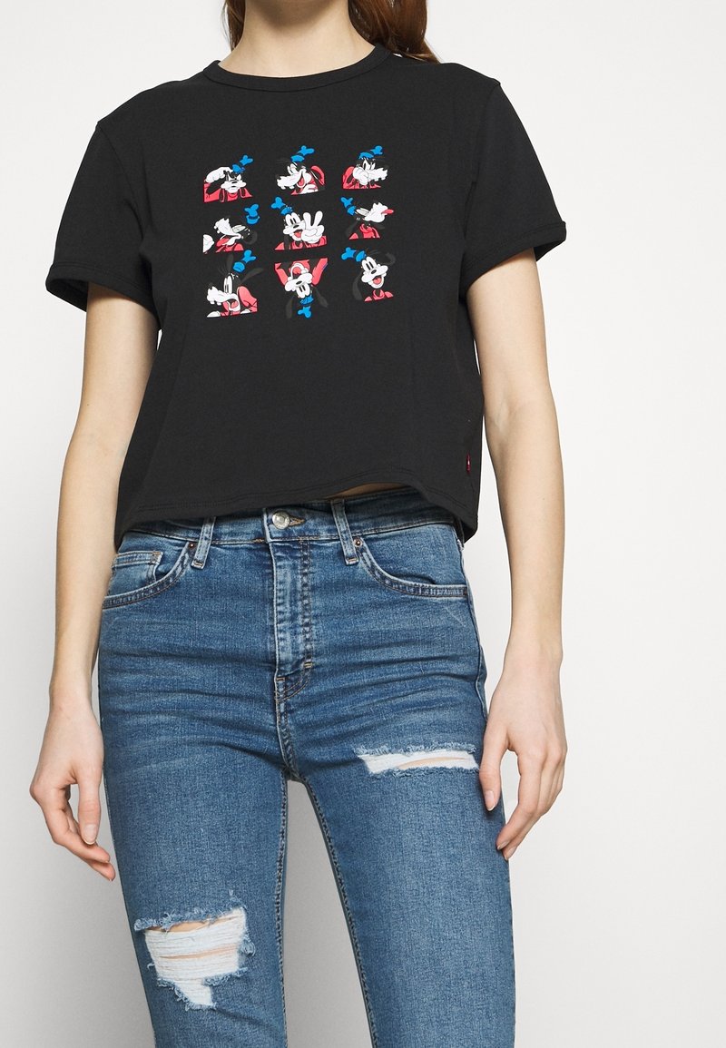 Personne portant un t-shirt court noir avec neuf visages de chiens de dessin animé colorés et un jean bleu déchiré taille haute.
