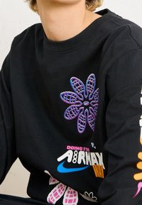 Svart långärmad sweatshirt med blommig grafik i lila och blått, med färgglada texter och logotyper på framsidan och ärmen. Mjukt tyg.