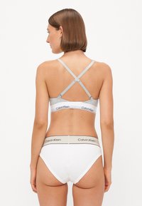 De beha heeft grijze stof met kruislings rugbanden en een witte tailleband met het label "Calvin Klein." De bijpassende slipjes zijn wit met een vergelijkbare tailleband.