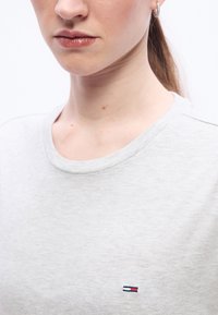 Κοντινό πλάνο ενός ατόμου που φοράει ανοιχτό γκρι πουκάμισο με ένα μικρό ορθογώνιο λογότυπο σε κόκκινο, άσπρο και μπλε στο στήθος και ένα σκουλαρίκι σε σχήμα κρίκου.