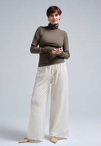 Brauner gerippter Rollkragenpullover, weiße weite Hose mit Kordelzug und hellfarbene spitze Schuhe. Glattes Material, lockere Passform.