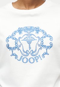 Λευκό πουκάμισο με μπλε περίτεχνο έμβλημα που απεικονίζει ένα στυλιζαρισμένο λουλούδι και διακοσμητικές μπούκλες, με τη λέξη "JOOP!" κάτω από το σχέδιο.