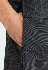 Crna tkanina s suptilnim valovitim uzorcima, koja ima bočnu džep sa detaljem cik-cak zatvarača, a ruka vidno umetnuta u džep.