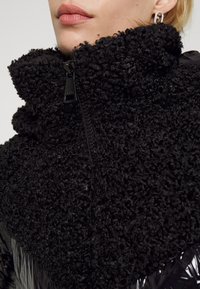 Svart ytterkläder med hög krage, som har en plushig, texturerad tyg på toppen och en slät, glänsande yta nedanför. Dragkedja fram.