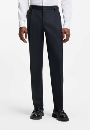 Mann trägt maßgeschneiderte schwarze Anzughose, weißes Hemd und schwarze Leder-Loafer, steht vor weißem Hintergrund.