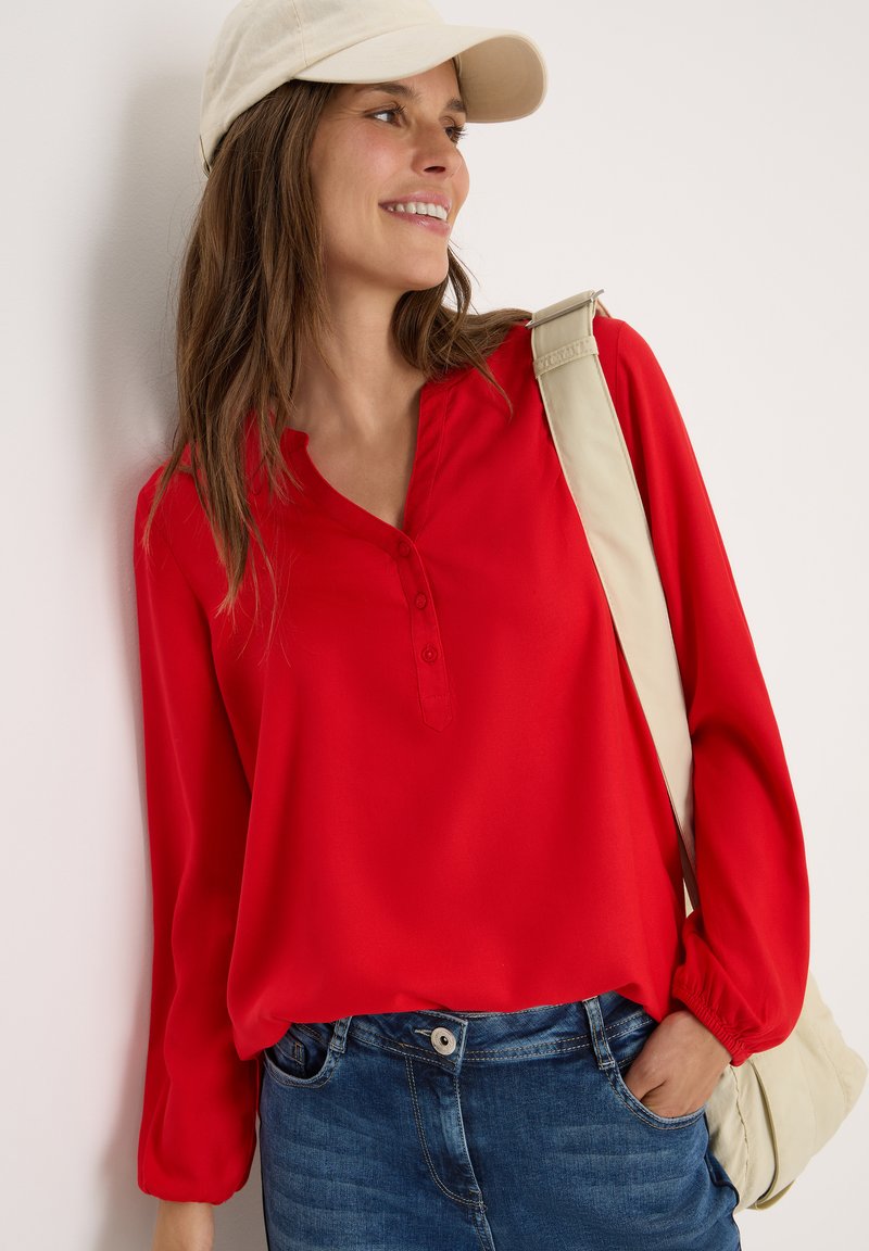 Blusa rossa a maniche lunghe con una patta di bottoni, in tessuto morbido e vestibilità rilassata, abbinata a jeans in denim blu e una borsa a spalla beige.