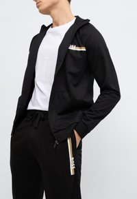Czarna bluza zapinana na zamek z kapturem, ozdobiona białymi i złotymi paskami na klatce piersiowej. Do kompletu czarne spodnie z logo i paskami po bokach.
