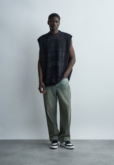 Ψηλός άνδρας που φορά αμάνικο μαύρο μπλουζάκι με σχέδιο, φαρδιά ξεβαμμένα πράσινα τζιν και ασπρόμαυρα αθλητικά παπούτσια, στέκεται μπροστά σε απλή φωτεινή τοίχο.