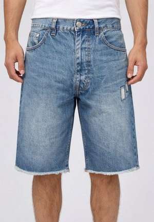 Pantaloncini di jeans blu con orli sfrangiati, leggero effetto consumato sulla coscia destra, indossati da una persona in piedi con le braccia rilassate.