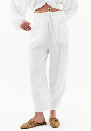Pantalons en lin légers et blancs avec une taille élastique et un cordon de serrage. Comprend des poches latérales et une coupe décontractée. Associés à des mules tressées couleur beige.