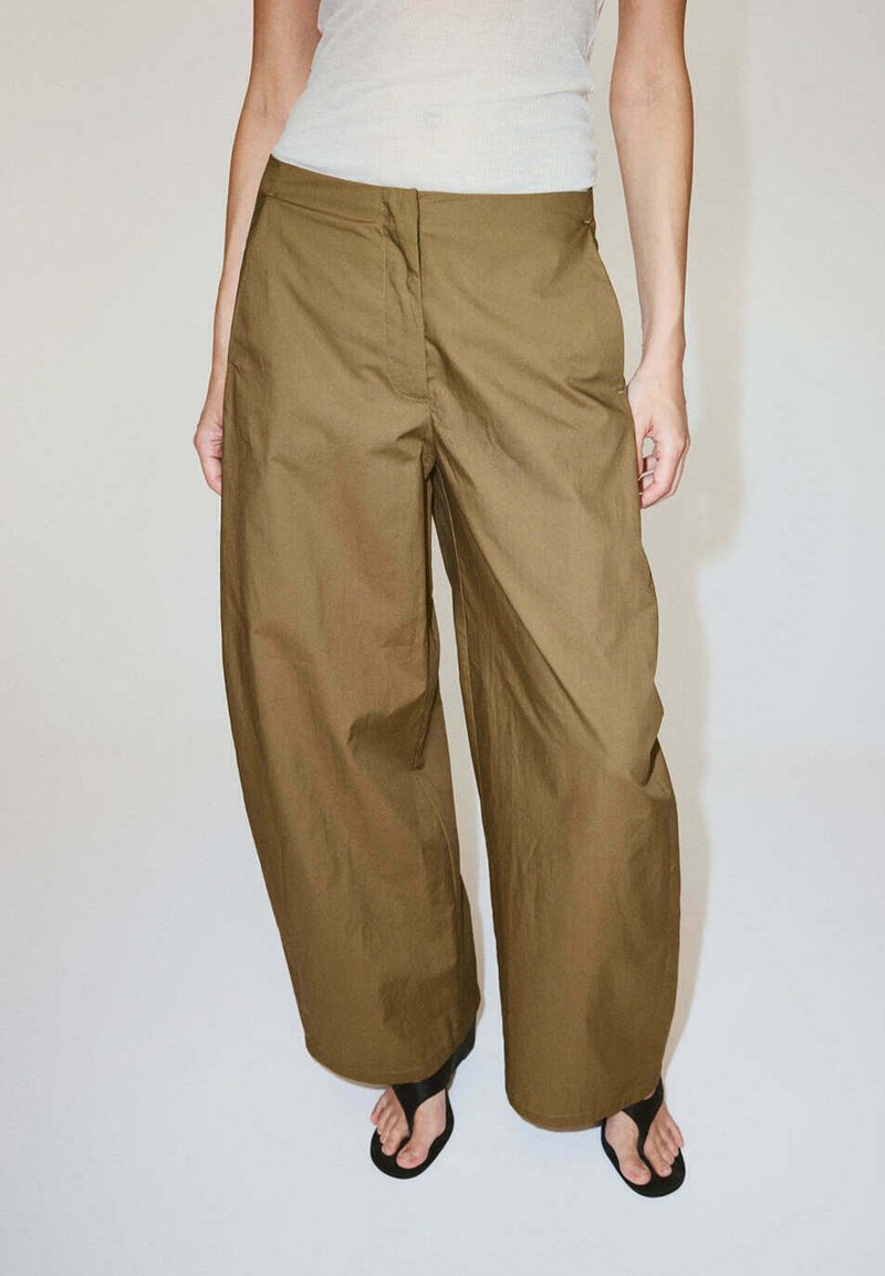 Personne portant un pantalon large vert olive associé à des tongs noires et un haut blanc sans manches sur un fond uni.