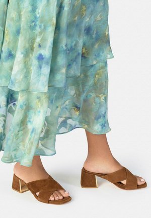 Pies con sandalias de tacón cuadrado de ante marrón con tiras cruzadas, combinadas con una falda ligera y transparente de capas con estampado floral en azul claro.