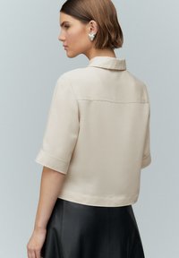 Beige blůza s krátkým rukávem a límcem, hladká textura a prošívané detaily na zadní straně, spárovaná s černou koženou sukní.
