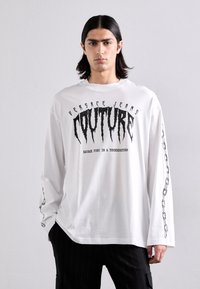 Balta garā piedurkne T-krekls ar melnu grafisko tekstu "COUTURE" un "SAVAGE FURY IN A THUNDERSTORM", ar ķēdes detaļām uz piedurknēm.