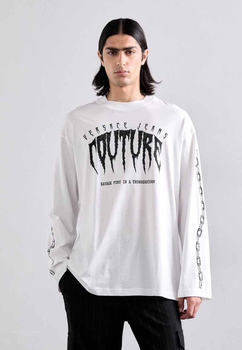 Balta garā piedurkne T-krekls ar melnu grafisko tekstu "COUTURE" un "SAVAGE FURY IN A THUNDERSTORM", ar ķēdes detaļām uz piedurknēm.