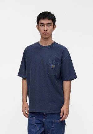 Mladý muž stojí čelom vpred, má na sebe tmavomodré oversized tričko s náprsným vreckom a modré džínsové nohavice.