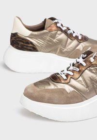 Zapatillas metálicas en dorado y marrón con acentos de ante, cordones blancos, patrón texturizado y una gruesa suela blanca. Presenta un detalle de estampado animal en el talón.